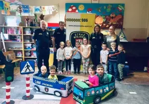 Zdjęcie grupowe dzieci ze Strażą Miejską. Z tyłu stoi baner Straży Miejskiej. Dzieci stoją na makiecie przejścia dla pieszych.