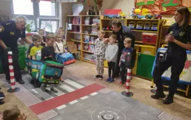 Dzieci w samochodach poruszają się po makiecie ulicy. Dw&oacute;ch chłopc&oacute;w i dziewczynka stoją na przejściu dla pieszych. Czekają na zielone światło.