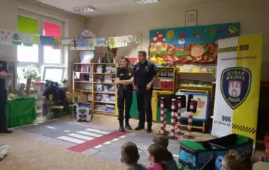 Strażnik i Strażniczka Miejska stoją na środku. Przed nimi na podłodze są rozłożone pasy drogowe. Obok nich stoją sygnalizatory świetlne. Dzieci siedzą przed nimi.