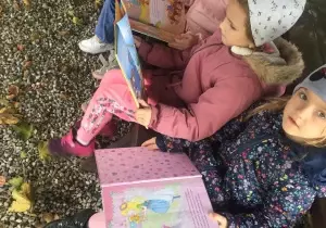 Dzieci siedzą na ławce. Czytają książki. Ubrane są w kurtki i czapki.
