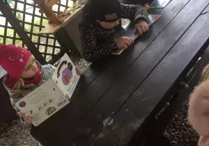 Trzy dziewczynki siedzą przy stoliku pod altaną. Czytaja książki.
