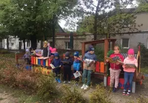 Dzieci stoją w ogrodzie przedszkolnym. W rękach trzyamją książki. Ubrazne są w czapki i kurtki.