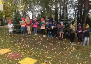 Dzieci stoją w lasku przedszkolnym. Trzymają w rękach książki. Czytają. Ubrazne są w kurtki i czpaki.