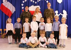 Zdjęcie grupowe. Dzieci są odświętnie ubrane. Razem z nimi stoją uczniowie ze szkoły wojskowej ubrani w mundury. 