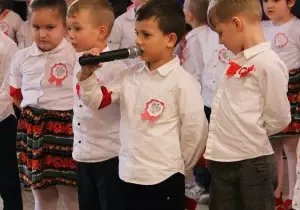 Dzieci odświętnie ubrane stoją na uroczystości. Chłopiec trzyma mikrofon. M&oacute;wi przez mikrofon.