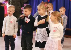 Dzieci odświętnie ubrane stoją na uroczystości. Chłopiec trzyma mikrofon. M&oacute;wi przez mikrofon.