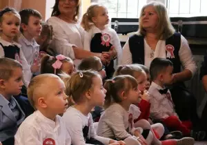 Dzieci i nauczycielka są odświętnie ubrani. Siedzą na uroczystości.