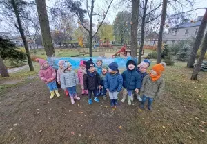 Zdjęcie grupowe dzieci w ogrodzie przedszkolnym. Dzieci ubrane są w kurtki i czapki. Za nimi jest strecz zawieszony o drzewa.