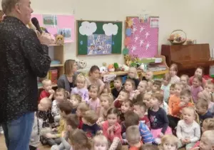 Dzieci siedzą na dywanie. Przed nimi stoi pan z mikrofonem. M&oacute;wi coś.