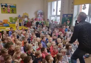 Dzieci siedzą na dywanie. Przed nimi stoi pan z mikrofonem. M&oacute;wi coś.