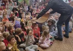 Dzieci siedzą na dywanie. Przed nimi stoi pan Witek i podaje zaczarowany dywan.