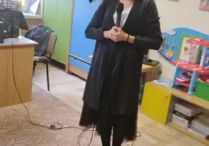 Pani trzyma mikrofon. Usmiecha się. Ma na sobie długi, czarny sweter.