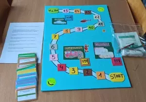 Na stole leży gra planszowa. Obok niej po lewej stronie instrukcja i kolorowe karty z pytaniami. Po prawej stronie folia z akcesoriami.