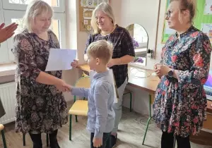 Pani Dyrektor trzyma za rękę chłopca. Gratuluje mu. W drugiej ręce trzyma dyplom. Z tyłu za chłopcem i obok chłopca stoi nauczycielka. Przygląda się.