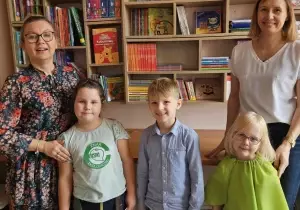 Tr&oacute;jka dzieci stoi na środku. Uśmiecha się. Po bokach stoją nauczycielki. Uśmiechają się do zdjęcia.