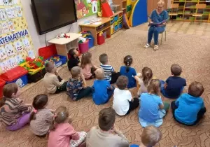  Dzieci siedzą na dywanie. Przed nimi na krzesełku siedzi pani. Czyta książkę.