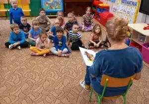 Dzieci siedzą na dywanie. Przed nimi siedzi pani. Trzyma książkę.