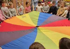 Dzieci stoją w kręgu. W środku jest kolorowa chusta animacyjna. Trzymają ją.
