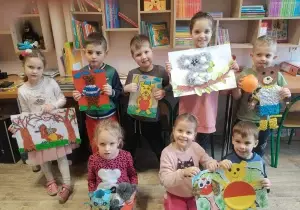 Zdjęcie grupowe dzieci. Dzieci stoją w bibliotece. Trzymają w rękach prace plastyczne misie wykonane na kartonach.