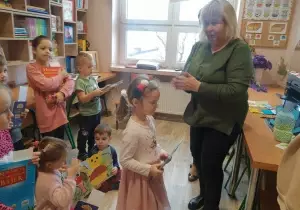 Pani dyrektor bije brawo. Dziewczynka stoi z książką. Pozostałe dzieci stoją z tyłu z książakami. Przyglądają się.