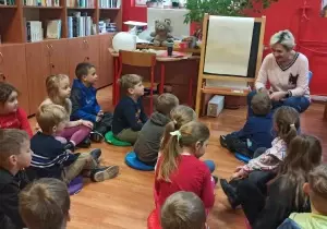 Dzieci siedzą w bibliotece na kolorowych pufach. Przed nimi siedzi na krześle bibliotekarka.
