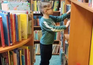 Chłopiec jest w bibliotece. Patrzy na p&oacute;łkę z książkami. Wyjmuje książkę z p&oacute;łki.