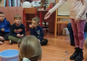 Dzieci siedzą w bibliotece na podłodze. Na środku stoi miska z wodą. Dziewczynka wskazuje ręką na miskę.