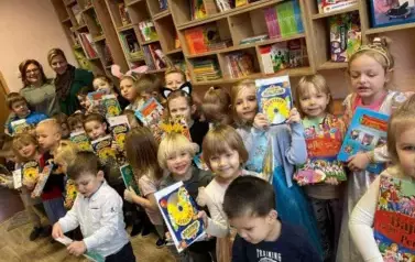 Dzieci stoją w bibliotece przedszkolnej. Uśmiechają się do zdjęcia. Trzymają w rękach książkę.