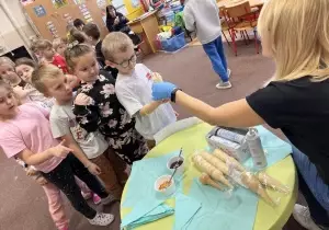 Dzieci stoją jeden za drugim w sali przedszkolnej. Nauczycielka siedzi przy stoliku. Wyciąga rękę z wafelkiem od lod&oacute;w do dziecka. 