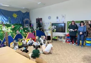 Dzieci siedzą na dywanie w sali przedszkolnej. Za nimi wisi dekoracja teatralna. W sali stoją nauczycielki z Czech.