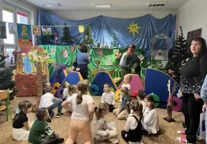Dzieci siedzą w kole w sali przedszkolnej. Za nimi wisi dekoracja teatralna. W sali stoją nauczycielki z Czech.