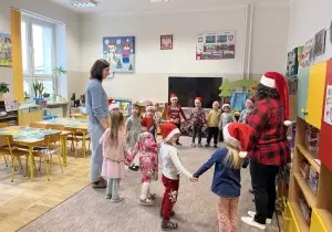 Dzieci wraz z nauczycielką i gościem z Czech stoją w kole w sali przedszkolenj. Nauczycielka ma czpakę mikołaja na głowie.