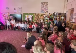 Goście siedzą w sali przedszkolnej na krzesełkach. Dzieci stoją na scenie.