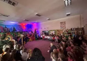 Goście siedzą w sali przedszkolnej na krzesełkach. Dzieci stoją na scenie.