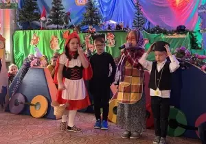 Dziewczynka przebrana za czerwonego kapturka stoi na środku. Obok chłopiec przebrany za wilka. Dalej dziewczynka przebrana za babcię m&oacute;wi przez mikrofon. Obok stoi chłopiec przebrany za zawiadowcę.