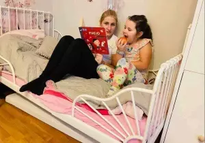 Mama czyta c&oacute;rce książkę. Dziewczynka gryzie jabłko. Siedzą w ł&oacute;żku w domu.