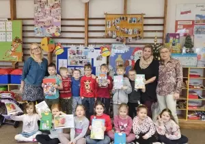 Niekt&oacute;re dzieci stoją. Trzymają w ręku książki. Niekt&oacute;re dzieci siedzą. Trzymają w ręku książki. Uśmiechają się do zdjęcia. Po prawej stronie stoją dwie nauczycielki. Po lewej stronie jedna nauczycielka.