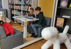 Dw&oacute;ch chłopc&oacute;w siedzi przy stoliku. Obok nich znajduje się regał z książkami.
