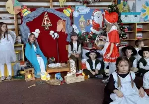 Zdjęcie szopki betlejemskiej w sali przedszkolnej. Przy szopce siedzi dziewczynka przebrana za Maryję i chłopiec przebrany za J&oacute;zefa. Przed szopką stoi dziewczynka przebrana za czerwonego kapturka.
