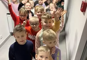 Dzieci uśmiechają się do zdjęcia. Machają. Stoją w parach jeden za drugim. Stoją w długim korytarzu.