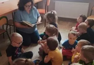 Nauczycielka z dziećmi siedzą w bibliotece przedszkolnej. Nauczycielka czyta dzieciom książkę. Dzieci słuchają.