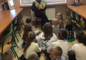 Nauczycielka z dziećmi siedzą w bibliotece przedszkolnej. Nauczycielka czyta dzieciom książkę. Dzieci słuchają.
