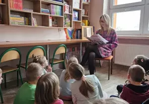 Nauczycielka z dziećmi siedzą w bibliotece przedszkolnej. Nauczycielka czyta dzieciom książkę. Dzieci słuchają.