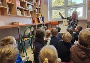 Nauczycielka z dziećmi siedzą w bibliotece przedszkolnej. Nauczycielka czyta dzieciom książkę. Dzieci słuchają.