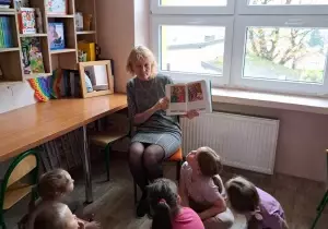 Nauczycielka z dziećmi siedzą w bibliotece przedszkolnej. Nauczycielka czyta dzieciom książkę. Dzieci słuchają.