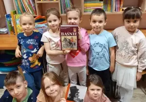 Dzieci stoją w grupie w bibliotece. Uśmiechają się do zdjęcia. Dziecko stojące na śrdoku trzyma książkę.
