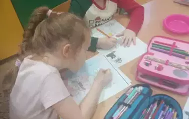 Dwie dziewczynki siedzą przy stoliku. Kolorują obrazki. Na stoliku leżą dwa pi&oacute;rniki.