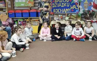 Dzieci siedzą na dywanie w p&oacute;kolu. Za nimi stoi nauczycielka. Za dziećmi wisi tablica z napisem babciu dziadku kochamy was.