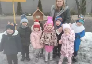 Zdjęcie grupowe dzieci w ogrodzie przedszkolnym. Dzieci ubrane są w kurtki i czapki. Uśmiechają się do zdjęcia. Razem z nimi stoi nauczycielka.