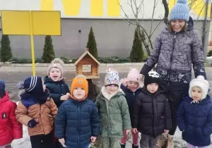 Zdjęcie grupowe dzieci w ogrodzie przedszkolnym. Dzieci ubrane są w kurtki i czapki. Uśmiechają się do zdjęcia. Razem z nimi stoi nauczycielka.